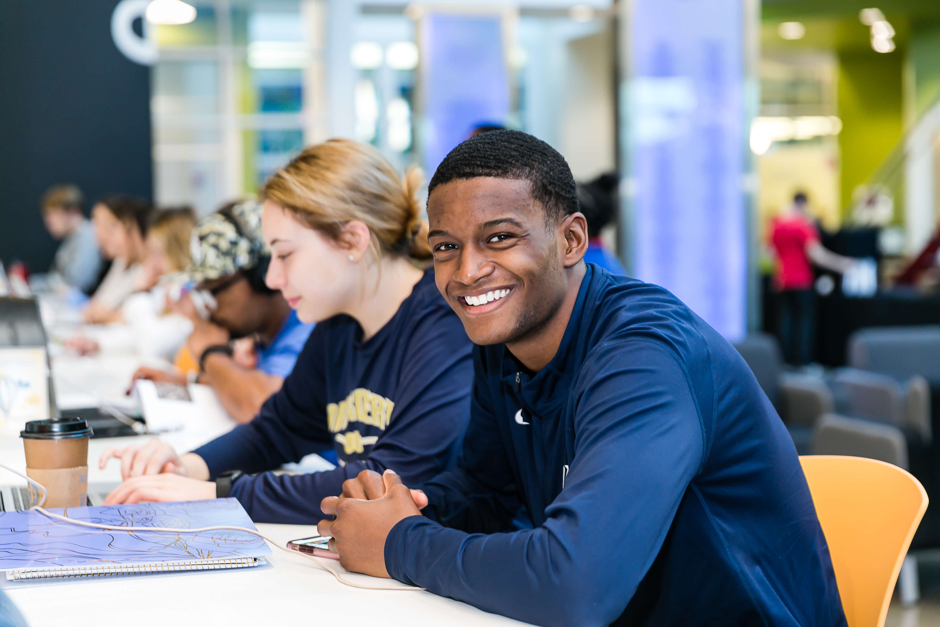 Smiling students working on laptops in a busy campus study area