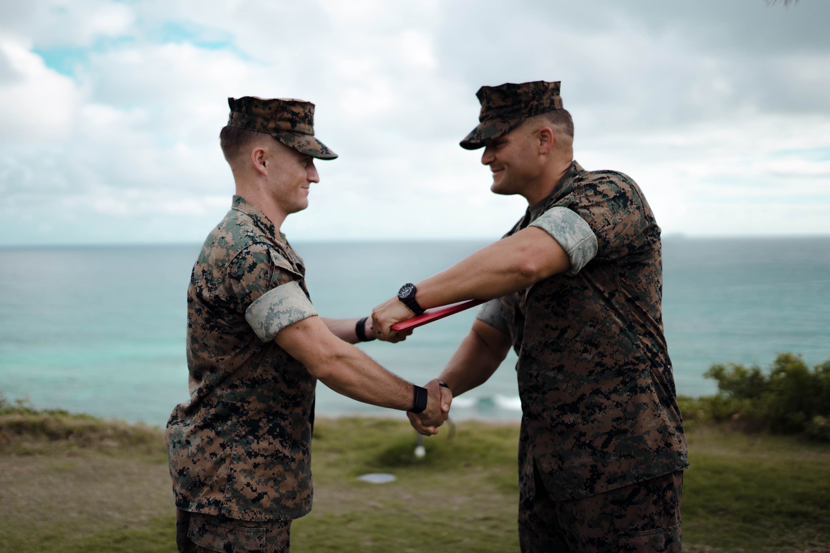 Two military service members in MARPAT camouflage uniforms share a handshake as one presents a red certificate folder to the other, set against an outdoor backdrop of green grass, the ocean, and a cloudy sky.
