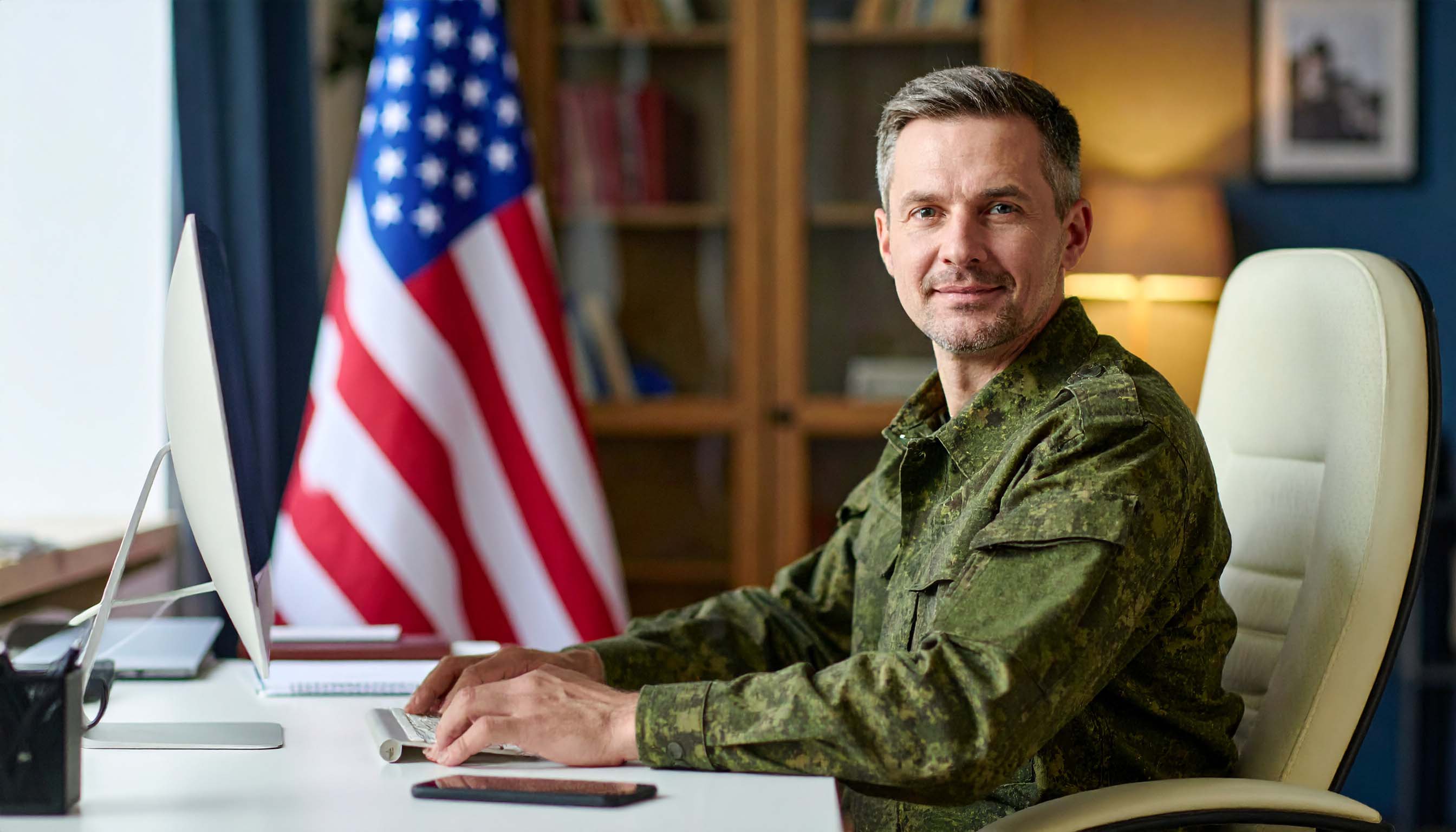 A confident military veteran in civilian clothes sitting at a laptop, studying or working on coursework, in a clean modern home office environment.