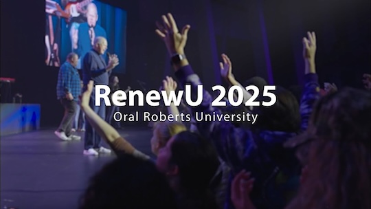 Video Thumbnail of a close up of the back of a few student's heads. One has his arms raised. In front of them you can see the praise and worship team on stage.